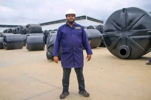 Man in purple work coat and hard hat standing at industrial facility with large metal storage tanks and aircraft in background