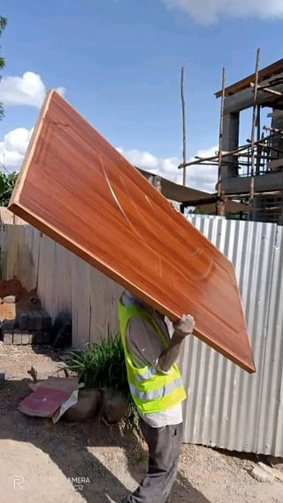 Construction worker in yellow safety vest carrying large rust-colored metal sheet at construction site with scaffolding in background