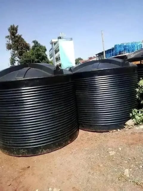 Two large black corrugated metal water storage tanks with domed covers in an outdoor industrial area with buildings visible in the background