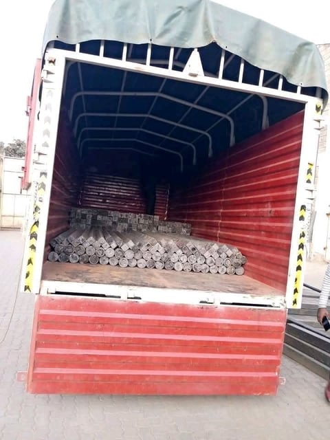 Open cargo truck loaded with stacked metal pipes and industrial materials on red flatbed with white railings