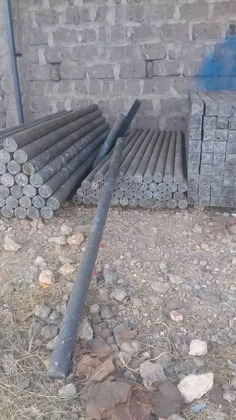 Bundles of dark metal rods and pipes stacked against a brick wall on rocky ground