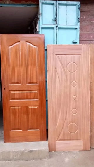 Three wooden doors leaning against a brick wall: a dark orange carved door, a light pink door with circular designs, and a blue-painted window frame behind them.