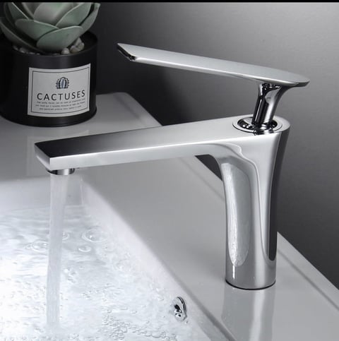 Modern chrome bathroom sink faucet with tall spout dispensing water into white basin, with potted cactus plant nearby