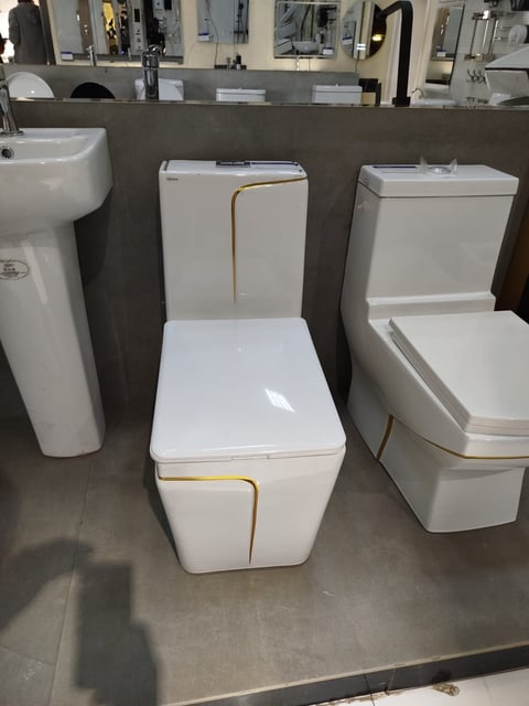 Three modern white toilets on display in a bathroom showroom with a pedestal sink visible on the left