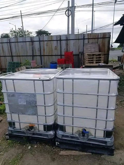 Two white plastic IBC totes with metal cages sit side by side on a dirt lot with corrugated metal fence and utility poles in background