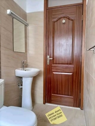 Bathroom interior with white pedestal sink, mirror, toilet, and wooden door with yellow business sign on tiled floor