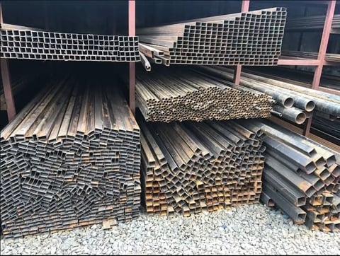 Industrial metal storage yard with stacks of square tubing, rectangular pipes, and steel channels organized in piles under a covered structure