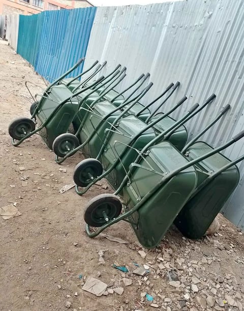 Row of green wheelbarrows lined up on dusty ground against corrugated metal fencing