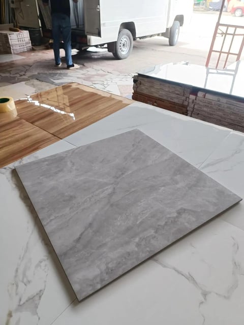 Gray concrete tile sample displayed on white marble flooring in a showroom with wood and dark samples visible in the background