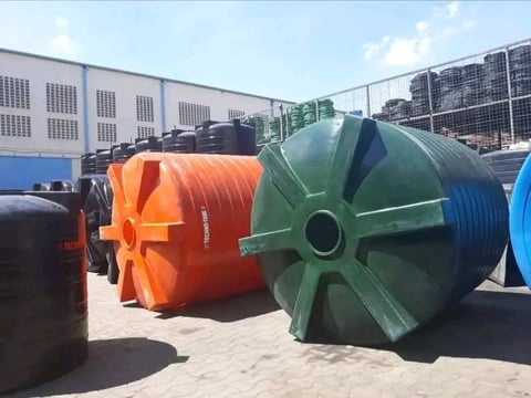 Industrial storage tanks in various colors (orange, green, blue, black) lined up outdoors at a facility with metal buildings in background
