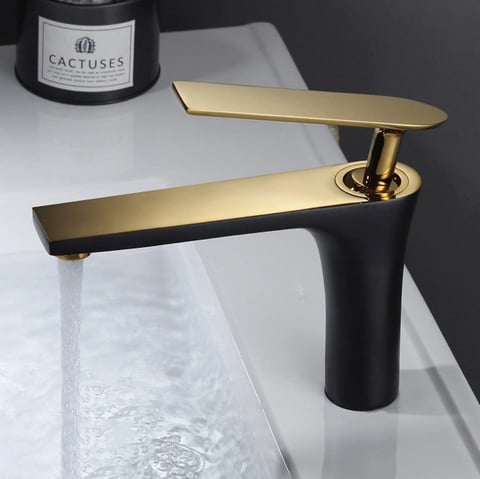 Modern gold and black bathroom faucet with rectangular spout over white sink, water running, styled with candle