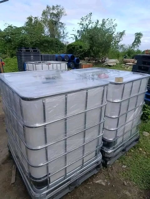 Two large plastic IBC tanks with metal cages stacked in an outdoor yard with grass and trees in the background
