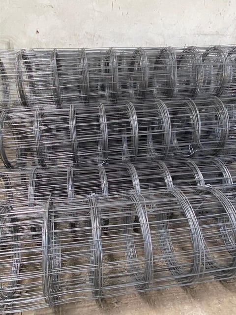 Stacked rolls of galvanized wire mesh fencing against a light-colored wall