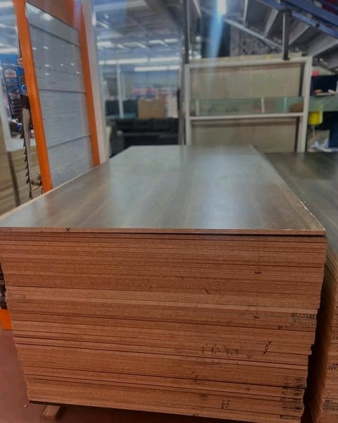 Stack of plywood sheets on a warehouse floor with metal shelving and orange structural elements in the background