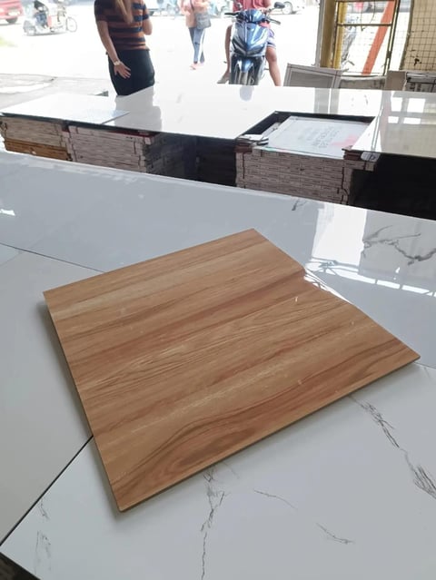 Wood grain sample tile displayed on white marble-look counter surface in retail showroom with people visible outside