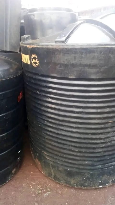 Large black cylindrical water storage tank with horizontal ridges and yellow emblem, positioned outdoors near other tanks