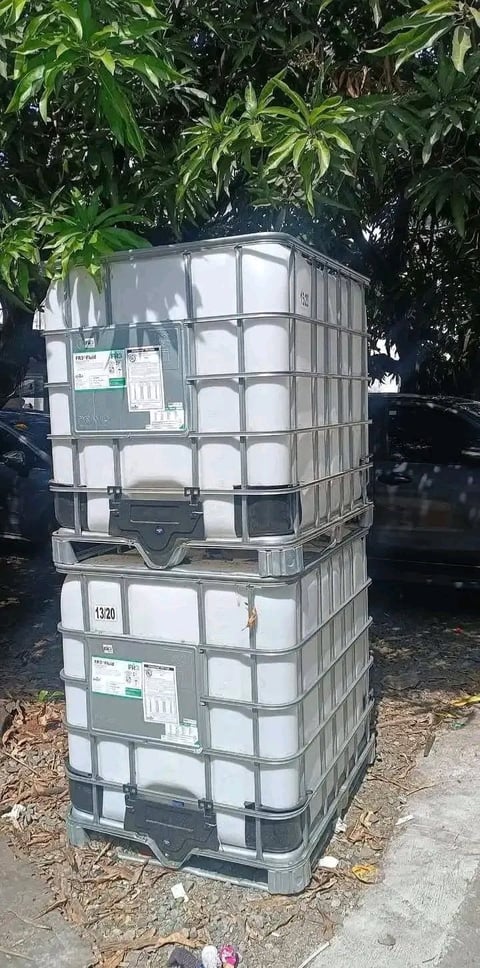 Stacked white plastic industrial storage containers with metal frames under green foliage
