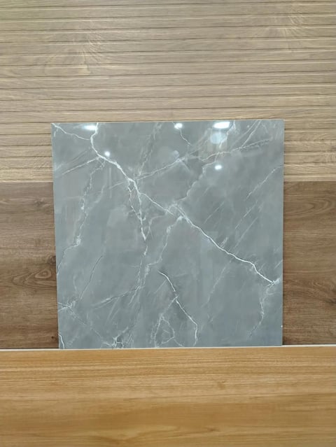 Gray marble tile sample with white veining displayed on a wooden floor against a wooden wall background