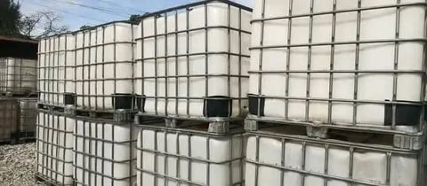 Row of large plastic storage tanks with metal cages and black valve connections in outdoor industrial setting