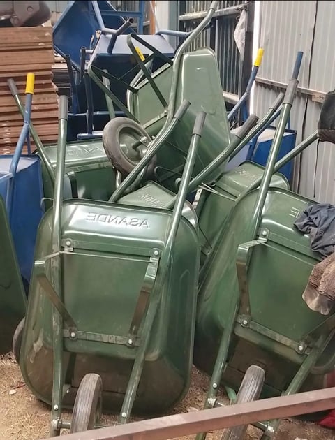 Green wheelie bins stacked on a cart in a yard with other equipment visible in the background